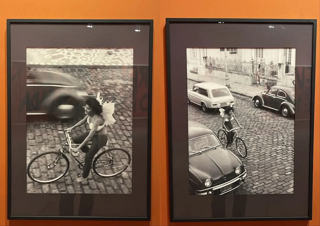 Montagem de duas obras de arte em preto e branco que mostram uma mulher andando de bicicleta trajando asas de anjo. Ambas estão colocados em uma parede laranja.