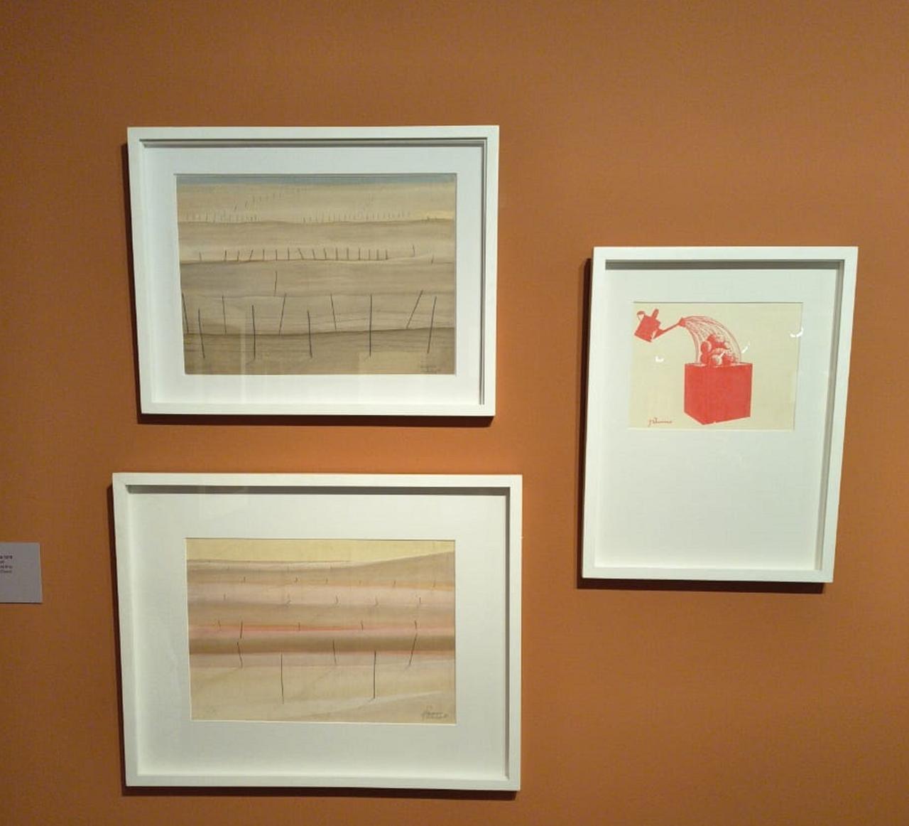 Três obras de Zé Tarcísio colocadas em uma parede laranja em um espaço expositivo.