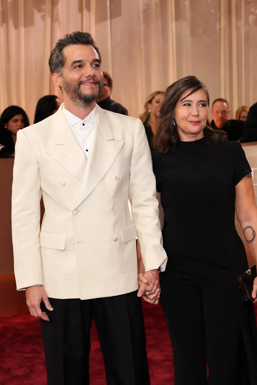Homem de terno branco e calça preta e mulher de vestido longo preto caminham de mãos dadas pelo tapete vermelho durante cerimônia de premiação, com fotógrafos, público e decoração elegante ao fundo.