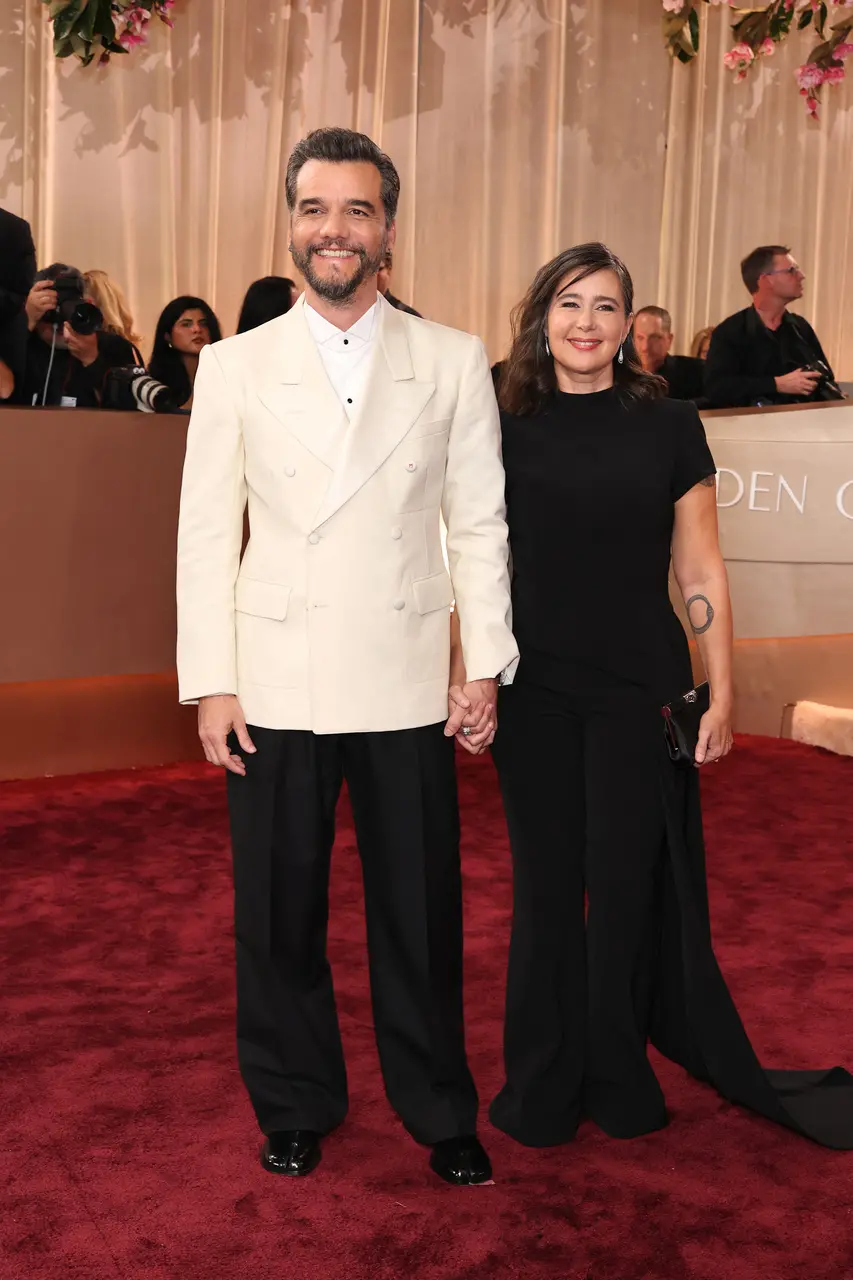 Homem de terno branco e calça preta e mulher de vestido longo preto caminham de mãos dadas pelo tapete vermelho durante cerimônia de premiação, com fotógrafos, público e decoração elegante ao fundo.