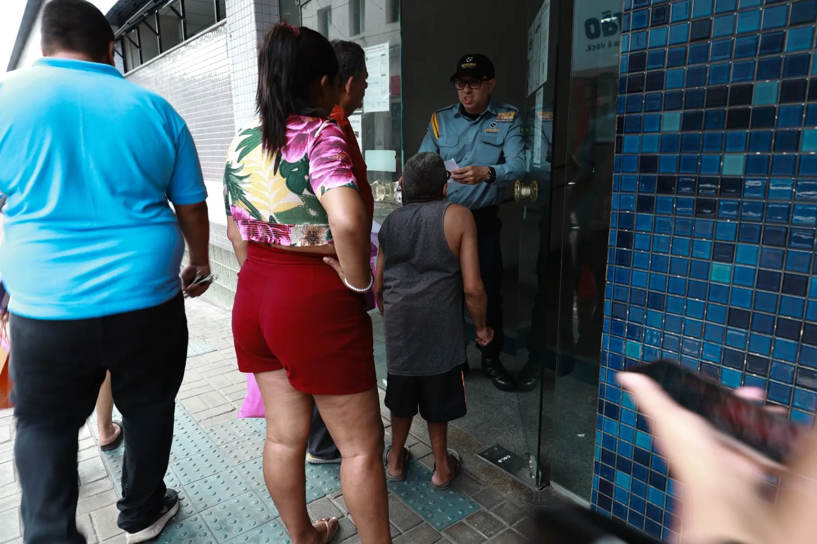 Pessoas aguardam em fila na entrada de uma agência, onde um segurança de uniforme azul e boné preto orienta um senhor na porta de vidro. Em primeiro plano, destacam-se as costas de um homem com camiseta azul e uma mulher vestindo shorts vermelhos e blusa floral. O ambiente possui paredes revestidas com pequenos azulejos azuis e piso tátil cinza.