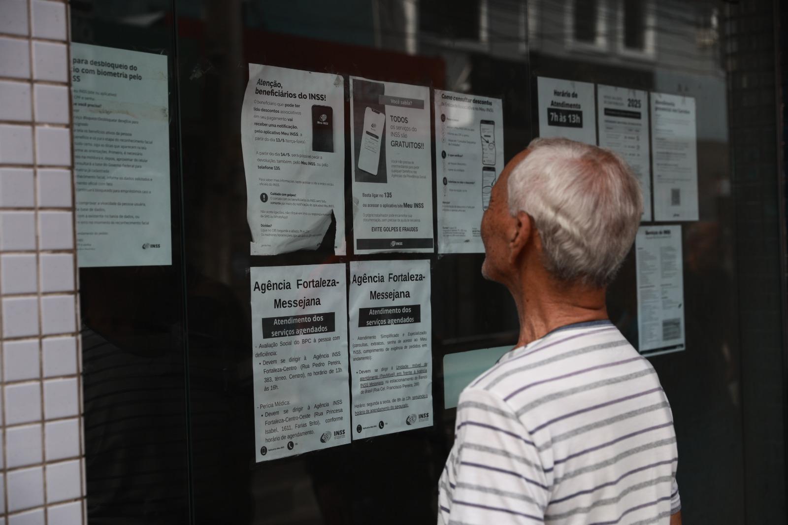 Um senhor de cabelos grisalhos e camisa listrada observa diversos cartazes informativos colados em uma porta de vidro. Os avisos contêm orientações sobre serviços do INSS, como o agendamento para a Agência Fortaleza-Messejana e alertas contra golpes. A cena ocorre em um ambiente externo com parede de azulejos claros ao lado da porta.