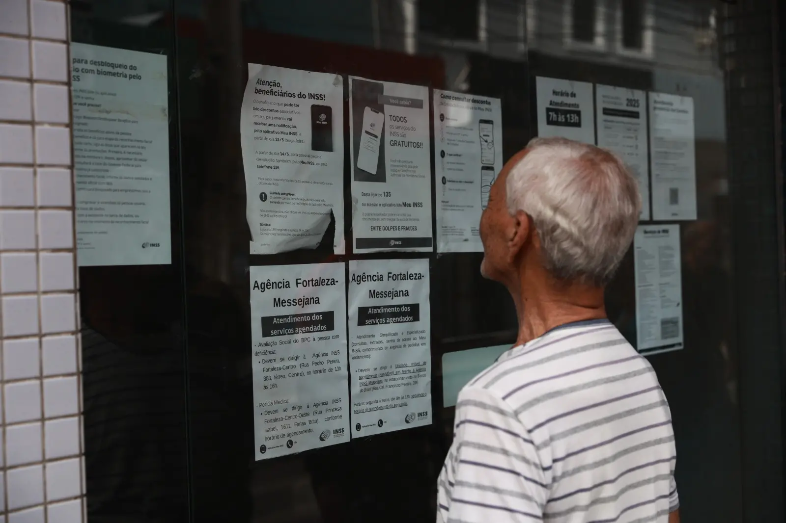 Um senhor de cabelos grisalhos e camisa listrada observa diversos cartazes informativos colados em uma porta de vidro. Os avisos contêm orientações sobre serviços do INSS, como o agendamento para a Agência Fortaleza-Messejana e alertas contra golpes. A cena ocorre em um ambiente externo com parede de azulejos claros ao lado da porta.