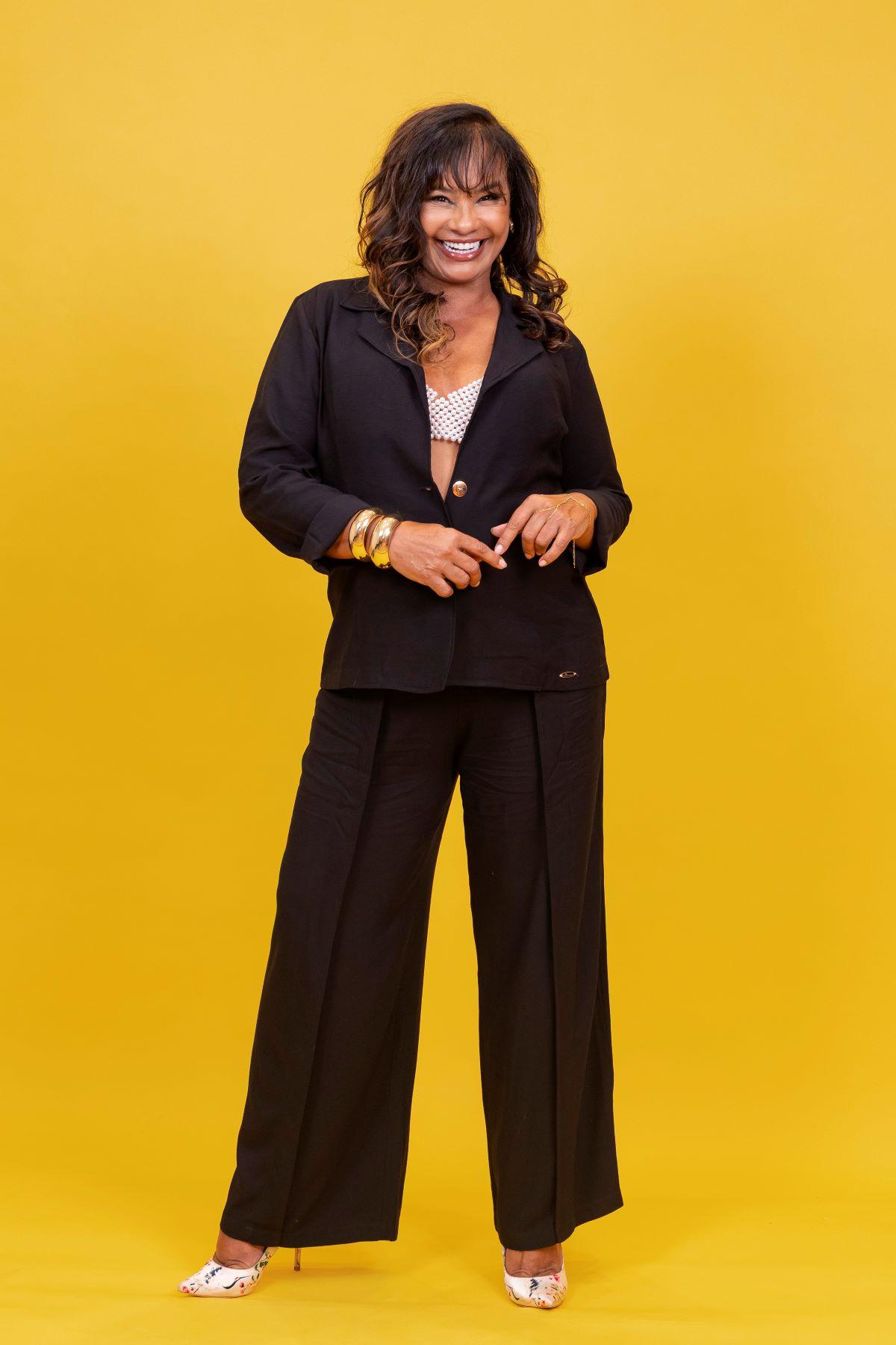 Retrato de corpo inteiro de uma mulher sorridente com cabelos longos, vestindo um blazer e calça social pretos, um top branco estampado e pulseiras douradas, em frente a um fundo amarelo vibrante. A mulher é Solange Couto.