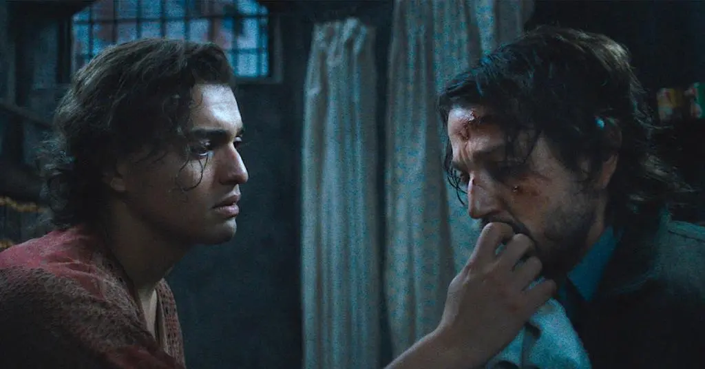 Os atores Tonatiuh e Diego Luna aparecem em um plano médio dentro de uma cela escura, onde Tonatiuh observa com preocupação e toca o rosto ferido de Luna. A iluminação fria e sombria destaca as texturas das roupas e as expressões de vulnerabilidade, compondo uma cena de forte carga dramática e melancolia.