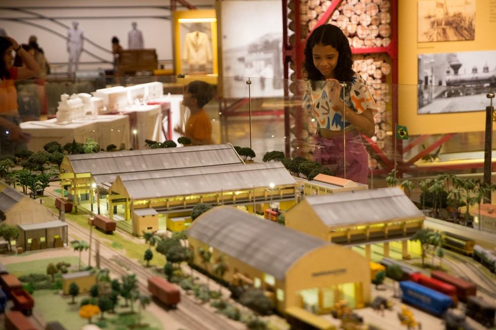 Na imagem, uma fotografia em plano médio e ângulo levemente elevado mostra o interior de um museu ferroviário. Em primeiro plano, há uma maquete detalhada de um pátio ferroviário, com galpões amarelos de telhados cinzas, pequenos vagões coloridos (vermelhos e azuis) sobre trilhos, árvores em miniatura e postes de luz acesos. Atrás de uma divisória de vidro que protege a maquete, uma menina jovem, de cabelos cacheados e negros, observa a miniatura com um sorriso gentil e olhar atento. Ela veste uma blusa branca com estampas coloridas e calças cor-de-rosa. O fundo da imagem está levemente desfocado, revelando outras exposições do museu, incluindo painéis informativos amarelos com fotografias históricas, uma pequena bandeira do Brasil e outras pessoas caminhando pelo espaço. A iluminação é quente e acolhedora, destacando os detalhes da maquete.