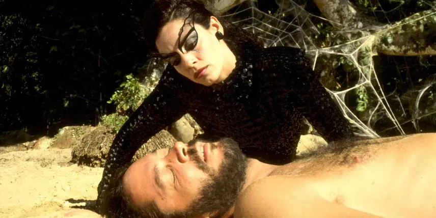 A atriz Sonia Braga, caracterizada com uma maquiagem artística de teia e traje preto brilhante, inclina-se sobre Raul Julia, que está deitado em uma praia ensolarada. O cenário ao fundo apresenta uma teia de aranha gigante entre as árvores, criando uma atmosfera onírica e dramática que mistura a natureza com elementos de fantasia.