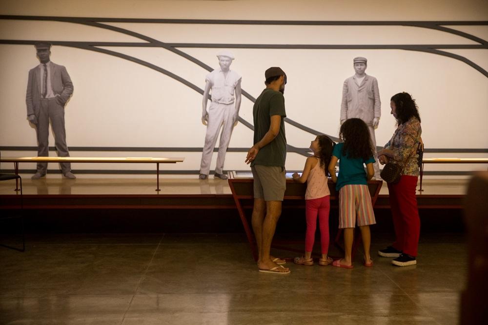 Na imagem, uma família composta por dois adultos e duas crianças observa uma exposição em um museu ferroviário. Ao fundo, uma parede branca exibe três figuras em tamanho real de trabalhadores ferroviários em preto e branco, posicionadas entre linhas pretas curvas que remetem a trilhos de trem. No centro, a família está reunida em torno de um painel interativo inclinado. O ambiente tem iluminação suave e piso de cimento queimado.