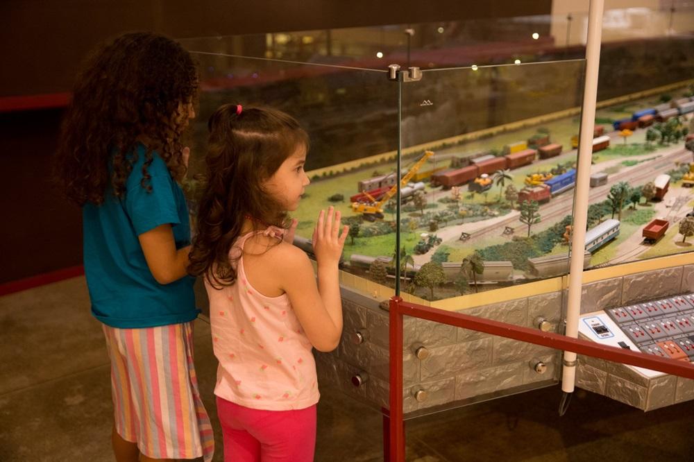 Na imagem, duas meninas, vistas de perfil, em pé diante de uma vitrine de vidro que protege uma maquete de ferreomodelismo. A menina à esquerda tem cabelos longos e cacheados e veste uma camiseta azul e saia listrada. A menina à direita tem cabelos castanhos ondulados presos em um meio-rabo e veste uma regata rosa clara e calça rosa choque. Ela está com as mãos encostadas no vidro, observando os detalhes. A maquete dentro da vitrine é vasta, apresentando diversos trens, vagões de carga coloridos, trilhos, miniaturas de árvores e guindastes amarelos. Na base da estrutura da maquete, há um painel de controle com diversos botões e interruptores vermelhos. A iluminação do ambiente é quente, criando uma atmosfera de curiosidade e encantamento.