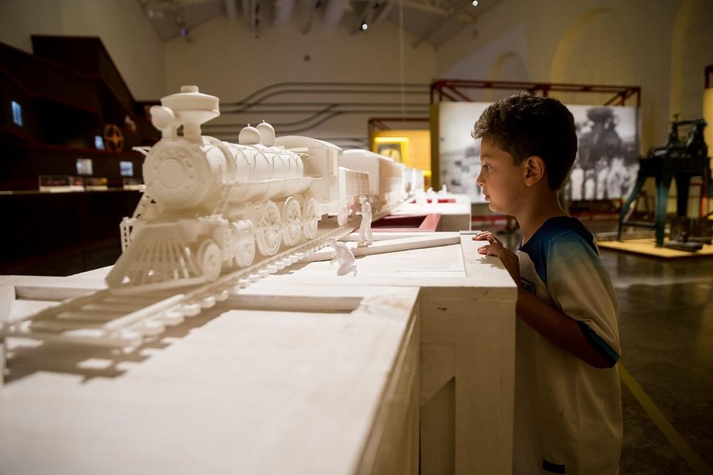 Na imagem, um menino de perfil observa atentamente uma grande maquete branca de um trem a vapor em um museu. A maquete, toda em tons de branco e detalhada, inclui a locomotiva sobre trilhos e pequenas figuras humanas também brancas. O menino veste uma camiseta azul e branca. Ao fundo, o ambiente do museu é amplo, com iluminação suave, outras exposições e fotos históricas em painéis.