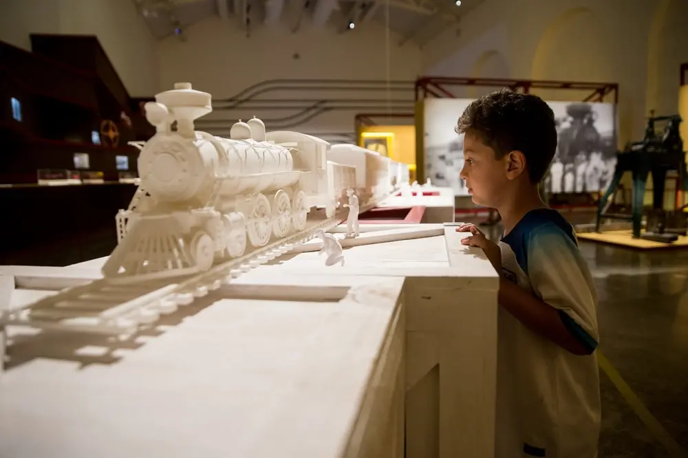 Na imagem, um menino de perfil observa atentamente uma grande maquete branca de um trem a vapor em um museu. A maquete, toda em tons de branco e detalhada, inclui a locomotiva sobre trilhos e pequenas figuras humanas também brancas. O menino veste uma camiseta azul e branca. Ao fundo, o ambiente do museu é amplo, com iluminação suave, outras exposições e fotos históricas em painéis.