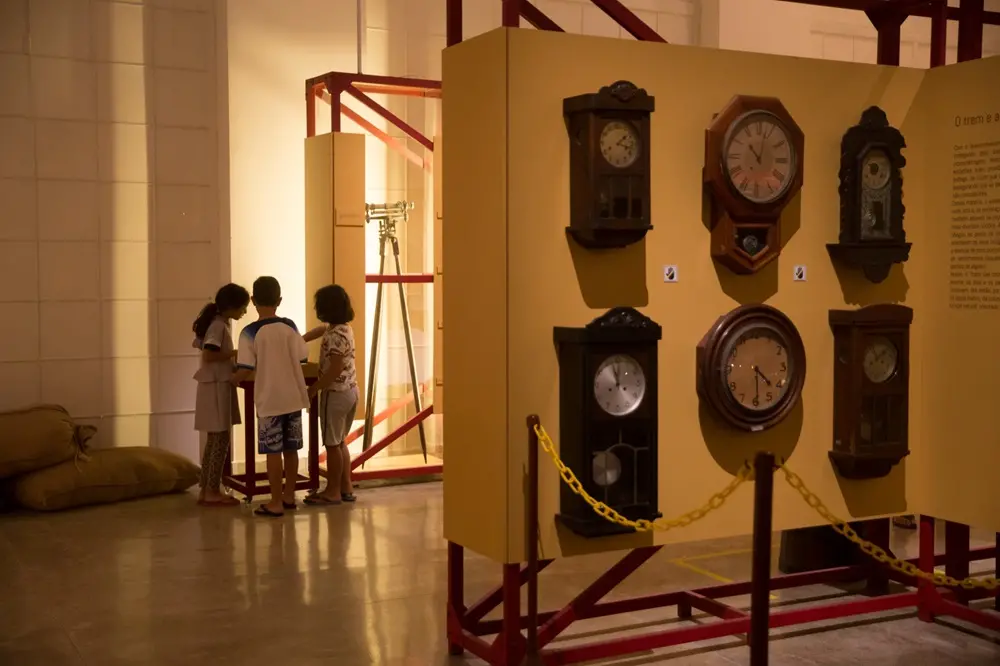 Na imagem, três crianças pequenas observam uma exposição no Museu Ferroviário. No primeiro plano, à direita, um painel amarelo exibe seis relógios de parede antigos de diferentes formatos e madeiras escuras. No centro da imagem, as crianças estão agrupadas em volta de uma peça interativa. Ao fundo, um teodolito (instrumento de medição) montado em um tripé está exposto dentro de uma estrutura metálica vermelha. A iluminação é suave e amarelada, destacando o ambiente histórico e educativo.