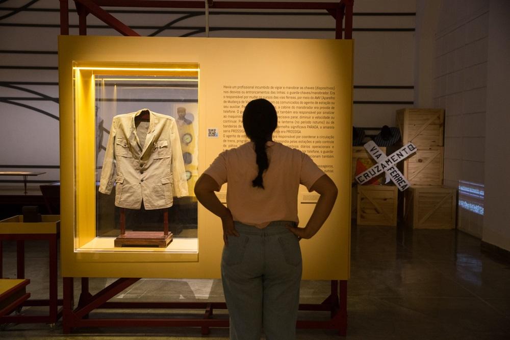 Na imagem, uma mulher vista de costas observa uma vitrine em um museu ferroviário. Ela tem cabelos escuros presos em uma trança e veste uma camiseta rosa clara e calça jeans. À sua frente, há um grande painel amarelo vibrante que contém: Uma vitrine iluminada à esquerda, exibindo uma túnica ou uniforme de época na cor bege, com botões frontais e bolsos. Um texto explicativo à direita, escrito em preto diretamente sobre o painel amarelo, detalhando as funções de profissionais ferroviários, como o guarda-chaves. Ao fundo, à direita, há caixotes de madeira empilhados e uma sinalização de trânsito ferroviária em formato de 