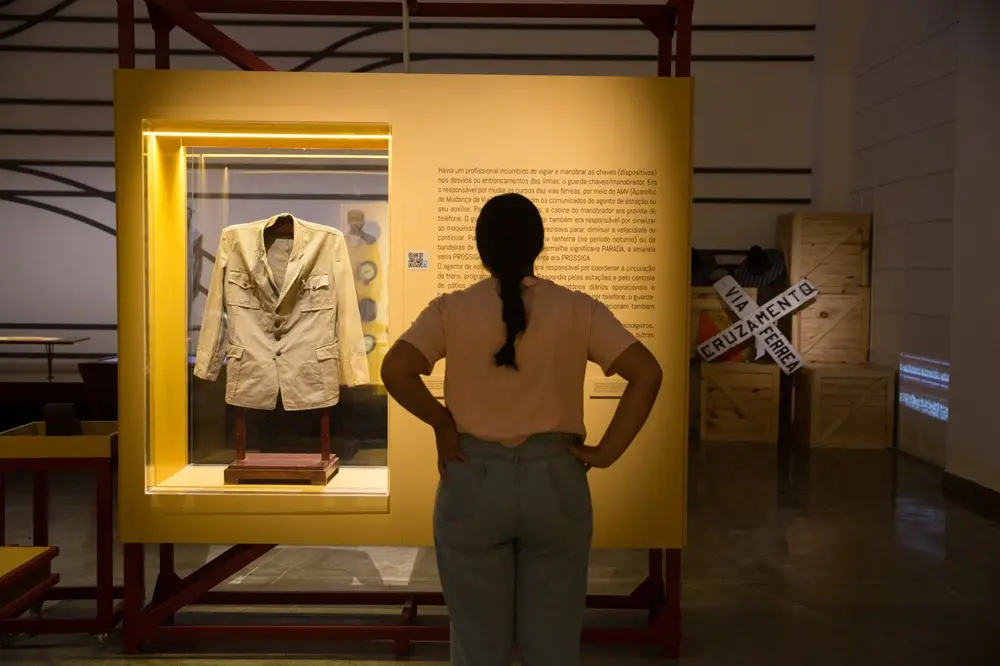 Na imagem, uma mulher vista de costas observa uma vitrine em um museu ferroviário. Ela tem cabelos escuros presos em uma trança e veste uma camiseta rosa clara e calça jeans. À sua frente, há um grande painel amarelo vibrante que contém: Uma vitrine iluminada à esquerda, exibindo uma túnica ou uniforme de época na cor bege, com botões frontais e bolsos. Um texto explicativo à direita, escrito em preto diretamente sobre o painel amarelo, detalhando as funções de profissionais ferroviários, como o guarda-chaves. Ao fundo, à direita, há caixotes de madeira empilhados e uma sinalização de trânsito ferroviária em formato de 