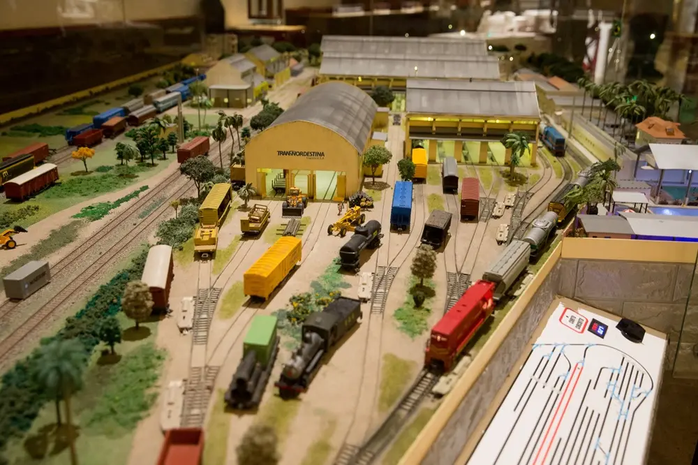 Na imagem, fotografia em ângulo elevado de uma maquete detalhada de um pátio ferroviário em um museu. No centro, destaca-se um galpão amarelo com o logotipo 