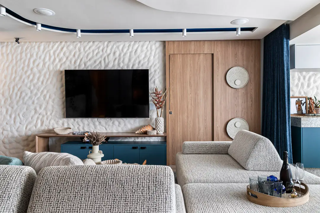 Uma parede branca com técnica de reboco grego ondulado serve como painel para uma TV, integrada a um rack azul e painéis de madeira clara que ocultam uma porta. O ambiente da sala de estar apresenta sofás em tons de cinza texturizado e detalhes em azul no teto e cortinas, criando uma atmosfera moderna e acolhedora.