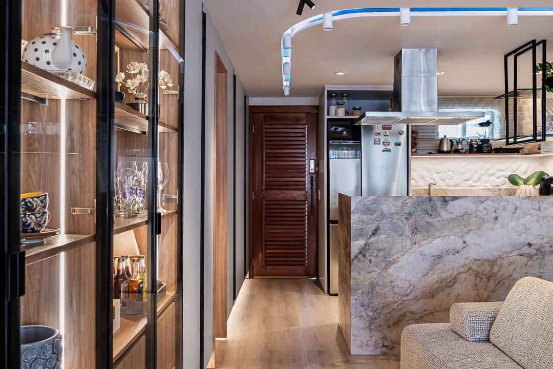 Uma cozinha integrada exibe uma bancada em mármore jaspeado que separa o ambiente da sala, destacando ao fundo uma parede com textura de reboco grego iluminada. O espaço é complementado por uma cristaleira elegante com portas de vidro e iluminação interna, além de detalhes em madeira e um rasgo de luz azul no teto que percorre o corredor.