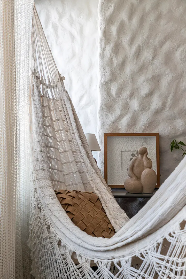 Uma rede de descanso branca com detalhes em macramê e uma almofada marrom ocupa o primeiro plano, transmitindo uma sensação de calmaria e serenidade. Ao fundo, uma parede com textura de reboco grego destaca uma escultura minimalista e um quadro decorativo, reforçando a estética de bem-estar do ambiente.
