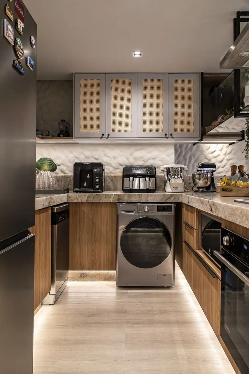 Uma cozinha funcional e iluminada apresenta armários superiores com palhinha indiana e armários inferiores em madeira, integrando eletrodomésticos modernos sob uma bancada de granito. A parede ao fundo exibe a textura de reboco grego realçada por iluminação embutida, criando um ambiente amplo e sereno que une praticidade e sofisticação.