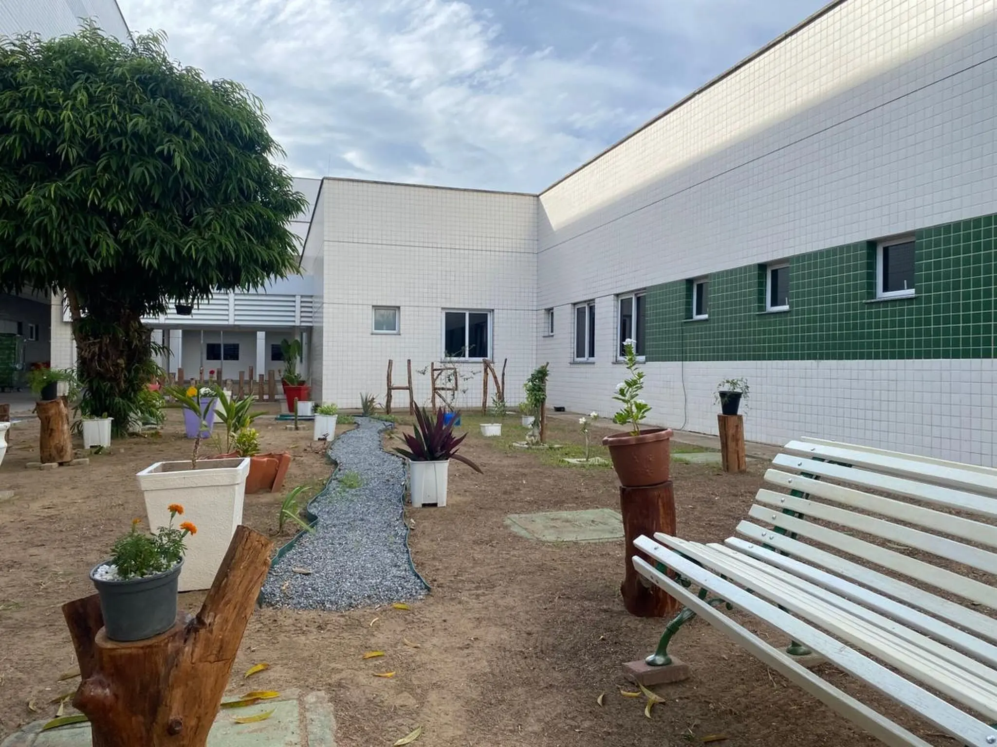 Jardim sensorial em pátio de hospital, com bancos, vasos de plantas e trilha de pedrinhas entre os canteiros.