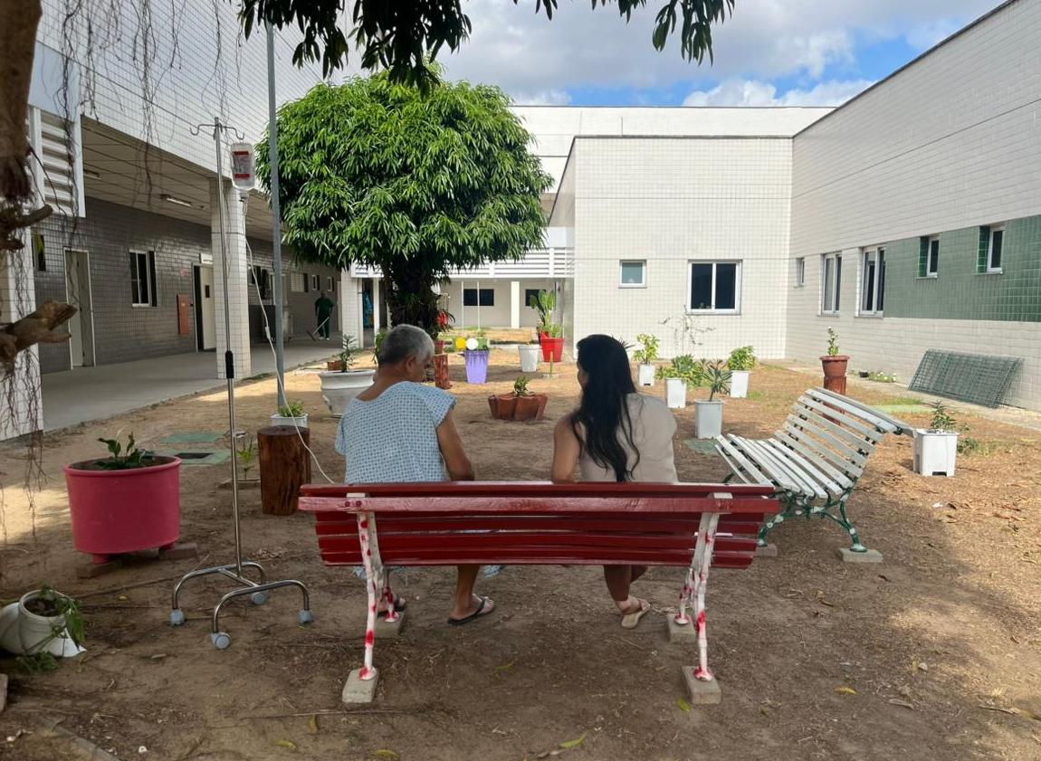 Duas pessoas sentadas em banco em jardim sensorial de hospital, com vasos de plantas ao redor.