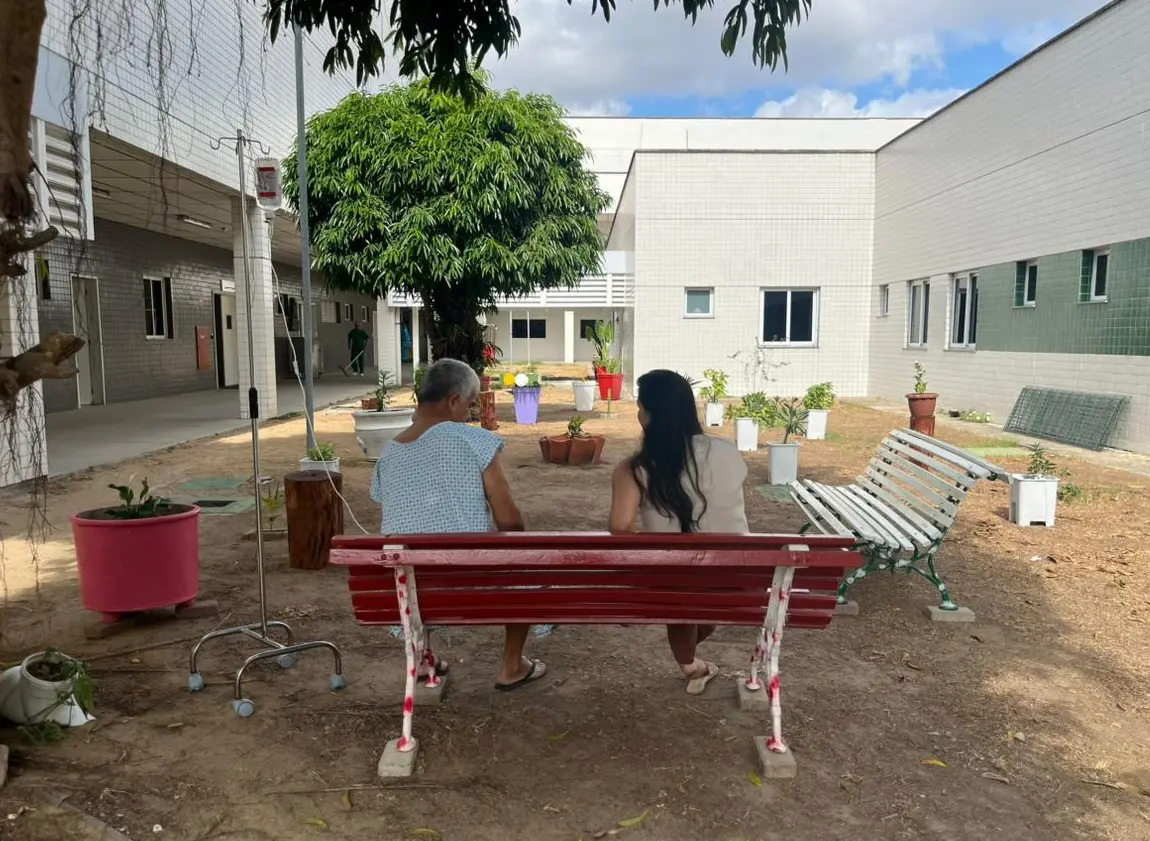 Duas pessoas sentadas em banco em jardim sensorial de hospital, com vasos de plantas ao redor.
