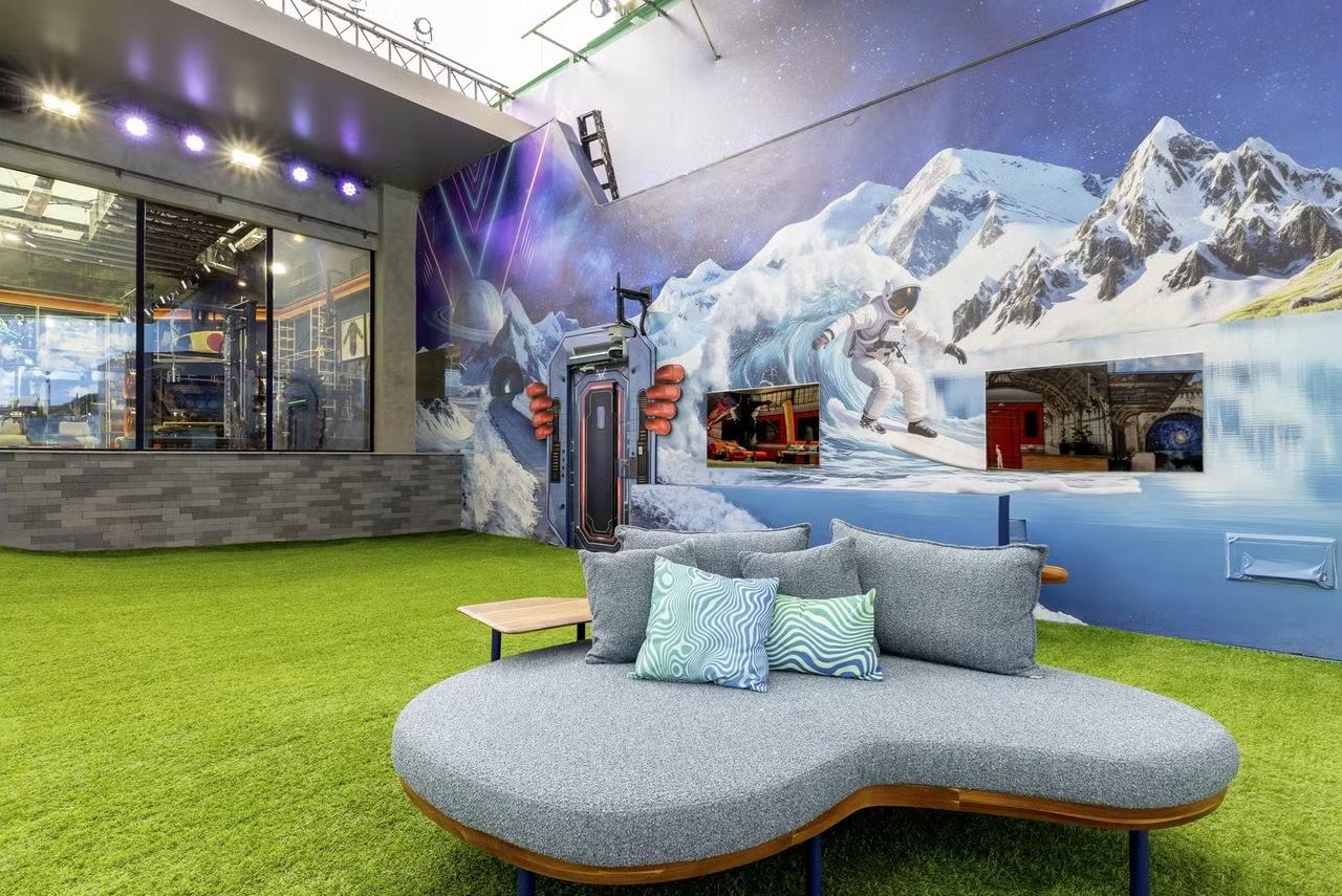 Um sofá cinza curvo com almofadas estampadas ocupa o centro de um jardim de grama sintética na área externa do BBB 26. Ao fundo, um painel artístico vibrante mistura montanhas nevadas e elementos espaciais, como um astronauta surfista, sob uma iluminação moderna e colorida.