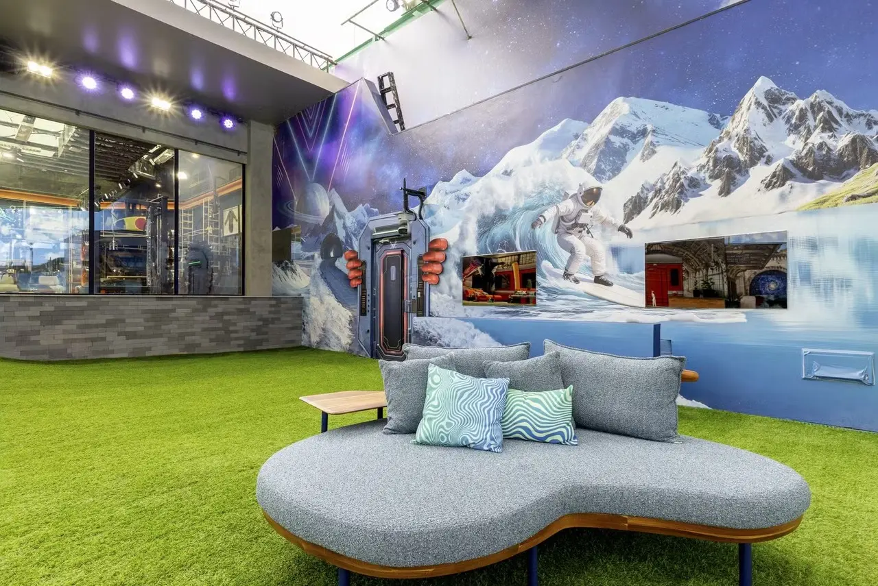 Um sofá cinza curvo com almofadas estampadas ocupa o centro de um jardim de grama sintética na área externa do BBB 26. Ao fundo, um painel artístico vibrante mistura montanhas nevadas e elementos espaciais, como um astronauta surfista, sob uma iluminação moderna e colorida.