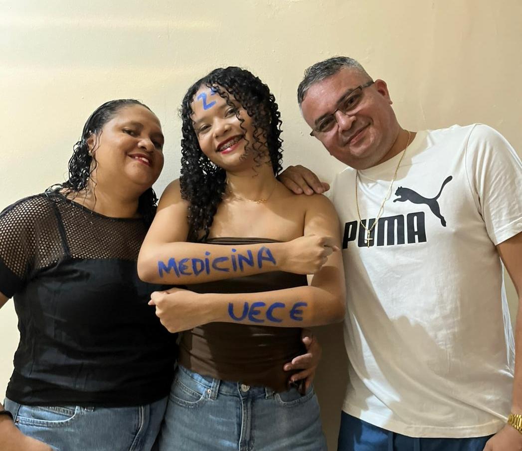 Ana Rafaely sorri ao lado dos pais, celebrando a aprovação em Medicina na UECE. Ela exibe os braços pintados com o nome do curso e da universidade em tinta azul.