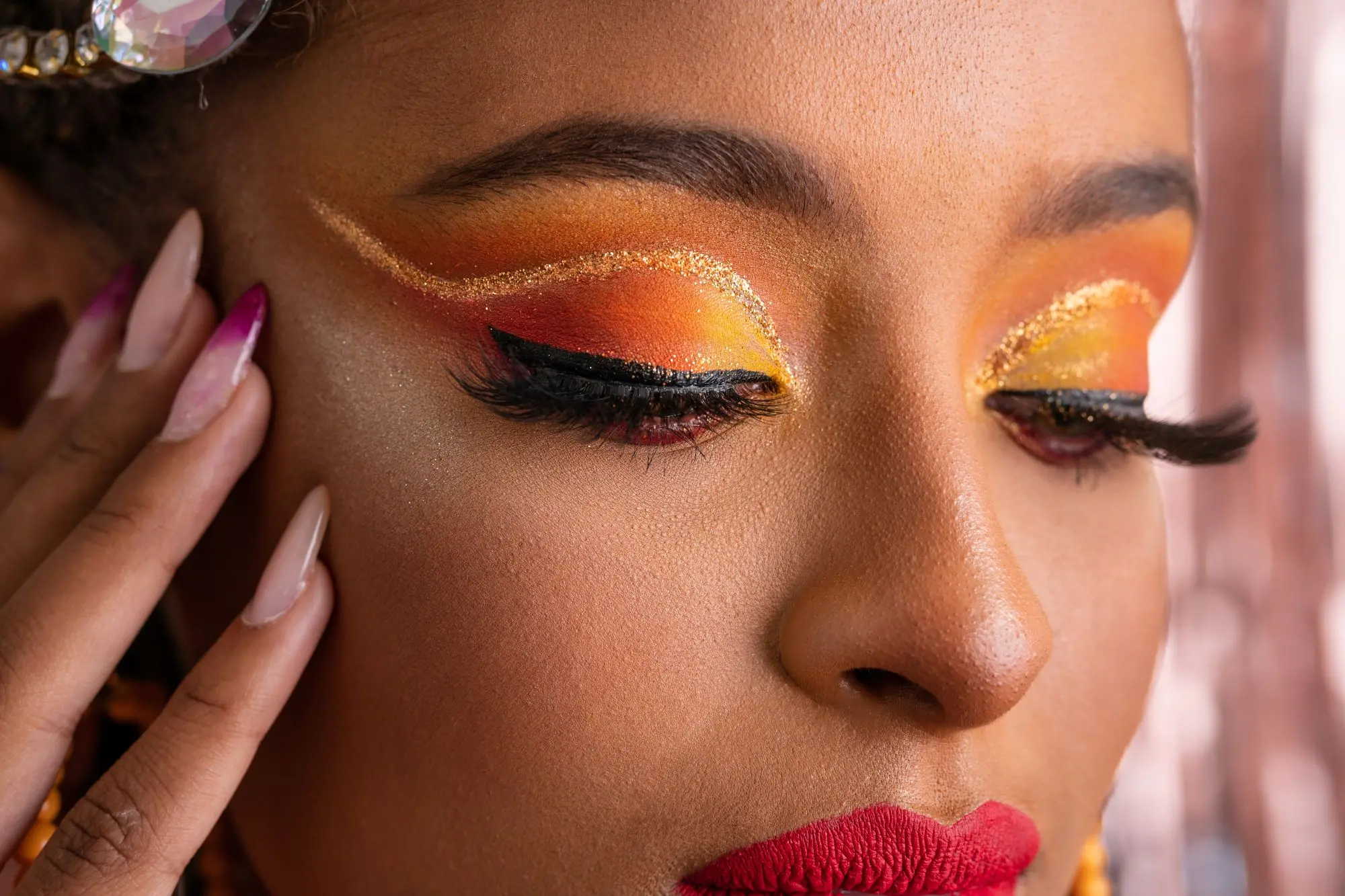 Close-up de uma maquiagem artística e vibrante com tons de laranja, amarelo e vermelho nas pálpebras, delineado dourado e cílios postiços volumosos. A pessoa tem batom vermelho e unhas longas.