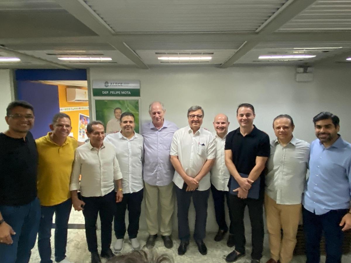 Grupo de dez homens em pé, lado a lado, posa para foto em ambiente interno iluminado por luz branca. Eles vestem roupas sociais e casuais, como camisas de botão e camisetas, e estão diante de uma parede com cartaz institucional ao fundo.