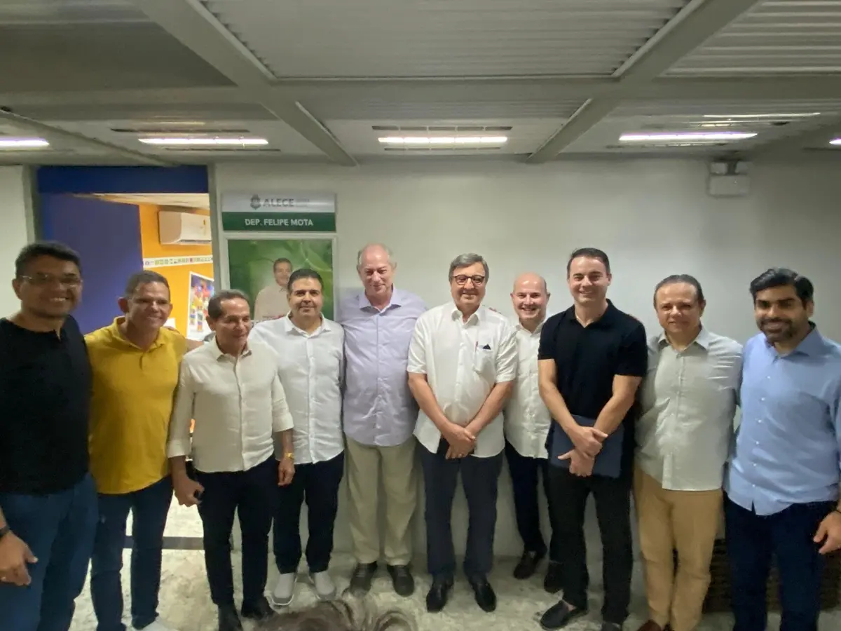 Grupo de dez homens em pé, lado a lado, posa para foto em ambiente interno iluminado por luz branca. Eles vestem roupas sociais e casuais, como camisas de botão e camisetas, e estão diante de uma parede com cartaz institucional ao fundo.