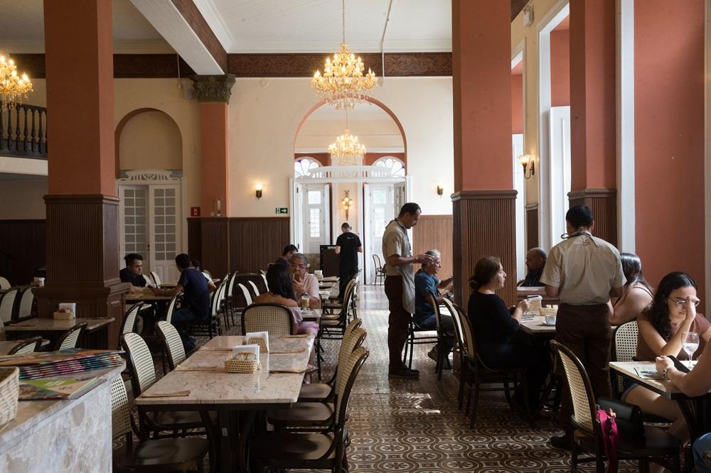 Na imagem, fotografia do salão interno de um café histórico. O ambiente possui grandes colunas em tons de terracota, teto branco com sancas decoradas e lustres de cristal pendentes. O chão é revestido com ladrilhos hidráulicos em tons de bege e marrom com padrões geométricos. Diversas mesas de mármore claro estão distribuídas pelo salão, acompanhadas por cadeiras de madeira escura com encosto de palhinha. Vários clientes estão sentados conversando ou consumindo, enquanto garçons vestidos com uniformes claros e aventais escuros circulam atendendo as mesas. Ao fundo, um arco emoldura a entrada para outra área iluminada, e grandes janelas laterais permitem a entrada de luz natural. O clima é de sofisticação acolhedora e preservação histórica.