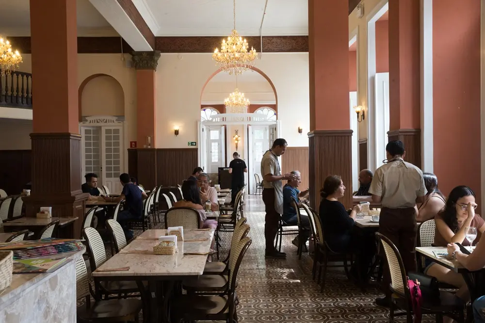 Na imagem, fotografia do salão interno de um café histórico. O ambiente possui grandes colunas em tons de terracota, teto branco com sancas decoradas e lustres de cristal pendentes. O chão é revestido com ladrilhos hidráulicos em tons de bege e marrom com padrões geométricos. Diversas mesas de mármore claro estão distribuídas pelo salão, acompanhadas por cadeiras de madeira escura com encosto de palhinha. Vários clientes estão sentados conversando ou consumindo, enquanto garçons vestidos com uniformes claros e aventais escuros circulam atendendo as mesas. Ao fundo, um arco emoldura a entrada para outra área iluminada, e grandes janelas laterais permitem a entrada de luz natural. O clima é de sofisticação acolhedora e preservação histórica.