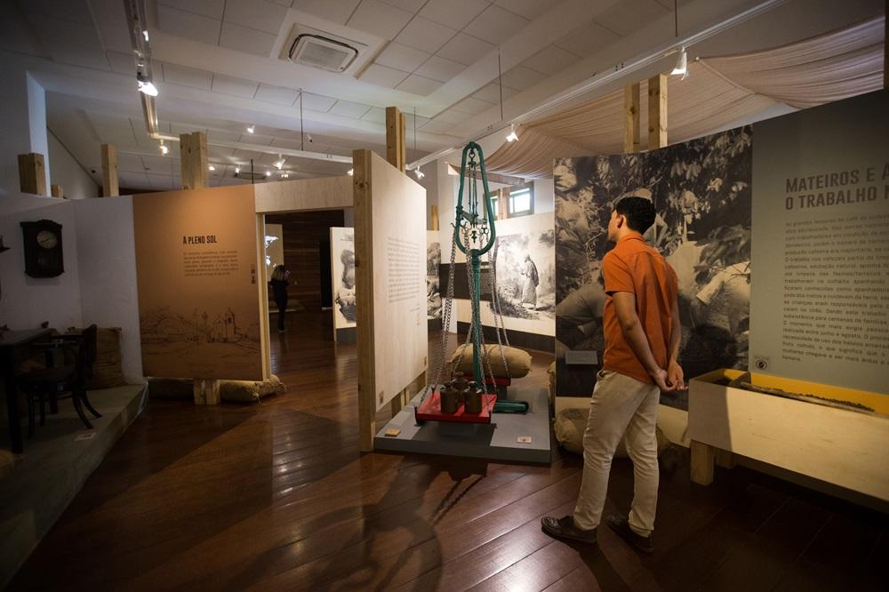 Na imagem, fotografia do interior de uma exposição no Museu da Indústria. Em primeiro plano, à direita, um homem jovem de costas, vestindo camisa laranja e calça clara, observa painéis informativos. No centro, há uma balança industrial antiga de metal verde, suspensa por correntes, com pesos de ferro sobre sua base de madeira. Ao redor, divisórias de madeira clara exibem textos e fotos históricas em preto e branco sobre a cultura do café. O ambiente tem iluminação suave e focada, teto branco com trilhos de luz e piso de madeira escura polida que reflete a claridade. Ao fundo, à esquerda, nota-se uma mesa de escritório antiga e sacos de estopa que remetem ao transporte de mercadorias.