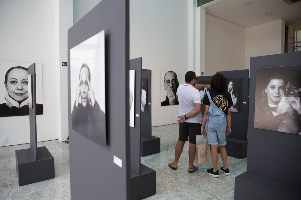 Na imagem, fotografia do interior de uma galeria de arte ampla e iluminada. Vários painéis cinza verticais estão espalhados pelo salão, exibindo retratos fotográficos em preto e branco de diferentes pessoas. No centro, um homem de camiseta branca e bermuda escura e uma jovem de macacão jeans e camiseta preta estão de costas, observando as obras. As paredes ao fundo são brancas e o chão é de granito claro.