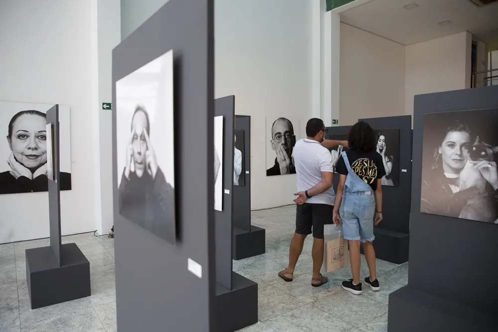 Na imagem, fotografia do interior de uma galeria de arte ampla e iluminada. Vários painéis cinza verticais estão espalhados pelo salão, exibindo retratos fotográficos em preto e branco de diferentes pessoas. No centro, um homem de camiseta branca e bermuda escura e uma jovem de macacão jeans e camiseta preta estão de costas, observando as obras. As paredes ao fundo são brancas e o chão é de granito claro.