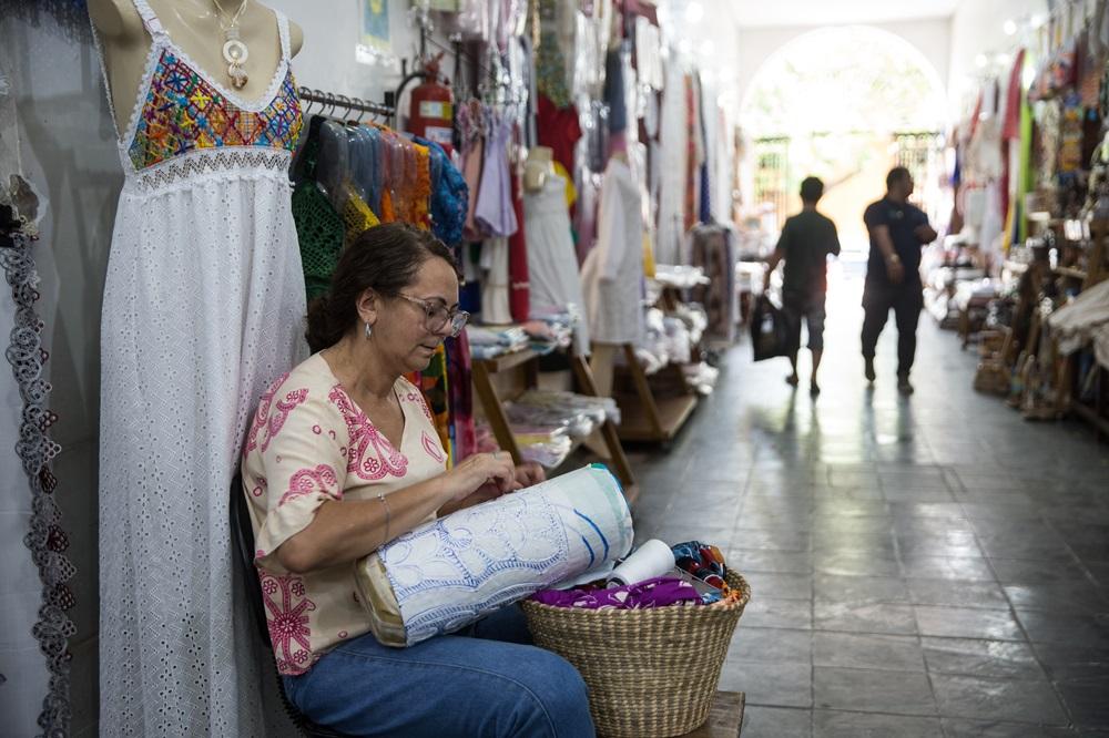 Na imagem, uma artesã sentada em um corredor de mercado de artesanato, trabalhando em uma renda de bilro sobre uma almofada cilíndrica apoiada no colo. Ela é uma mulher de pele clara, cabelos castanhos presos e usa óculos. À sua esquerda, há um manequim vestindo um longo vestido branco de renda. O corredor é ladeado por lojas repletas de roupas e redes coloridas, com dois homens caminhando ao fundo em direção a uma saída iluminada.