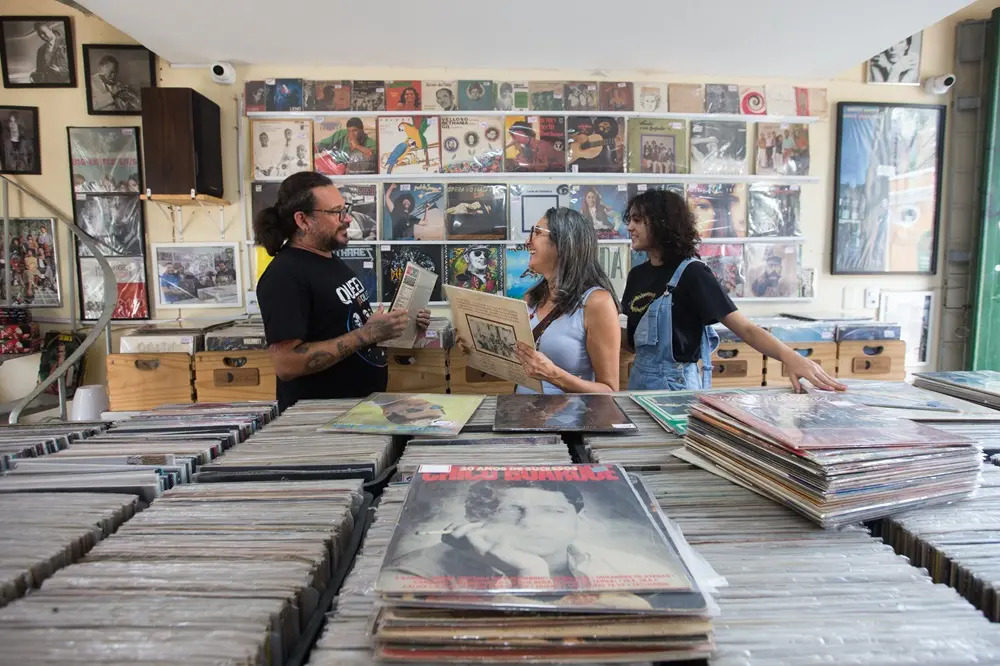 Na imagem, fotografia interna de uma loja de discos de vinil repleta de álbuns. No centro, três pessoas — um homem de camiseta preta à esquerda e duas mulheres à direita — conversam sorridentes enquanto seguram e examinam capas de LPs. O primeiro plano é dominado por grandes caixas e prateleiras horizontais cheias de discos, com destaque para um álbum de Chico Buarque na frente. Ao fundo, as paredes estão cobertas por dezenas de discos de vinil expostos em prateleiras, incluindo artistas da música brasileira, criando um ambiente cultural e nostálgico.