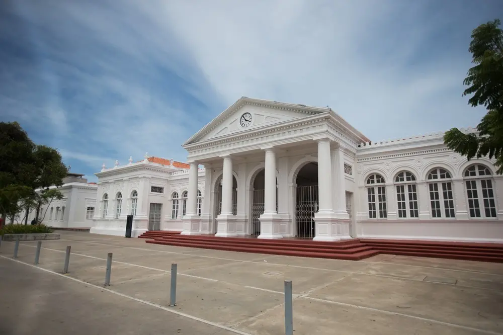Na imagem, fotografia colorida em ângulo levemente inferior da Estação das Artes. O edifício é totalmente branco e possui uma arquitetura clássica imponente. No centro, há um pórtico sustentado por quatro colunas robustas que terminam em um frontão triangular com um relógio circular. As laterais apresentam janelas altas em arco e detalhes ornamentais na platibanda. O chão à frente é de concreto claro com pequenos pilares metálicos de sinalização (balizadores) e degraus terracota que levam à entrada. O céu ao fundo é azul com nuvens suaves e há folhagens verdes nas extremidades laterais da foto.