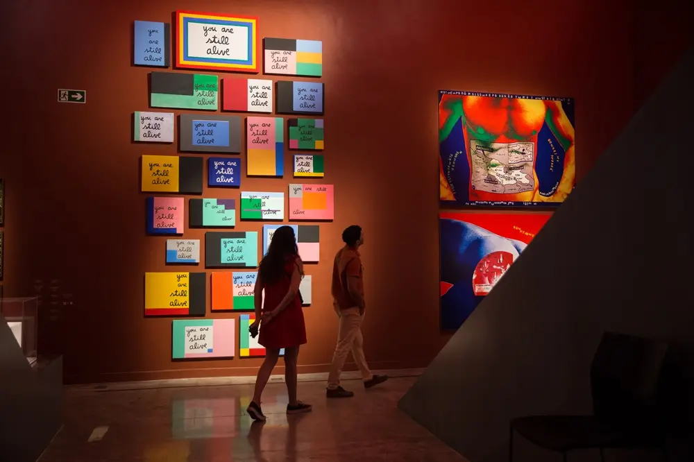 Na imagem, fotografia colorida do interior da Pinacoteca do Ceará. Em uma parede de tom terracota escuro, destaca-se uma instalação artística composta por diversas telas retangulares de tamanhos variados, dispostas verticalmente. Cada tela apresenta cores vibrantes e blocos geométricos, contendo a frase repetida 