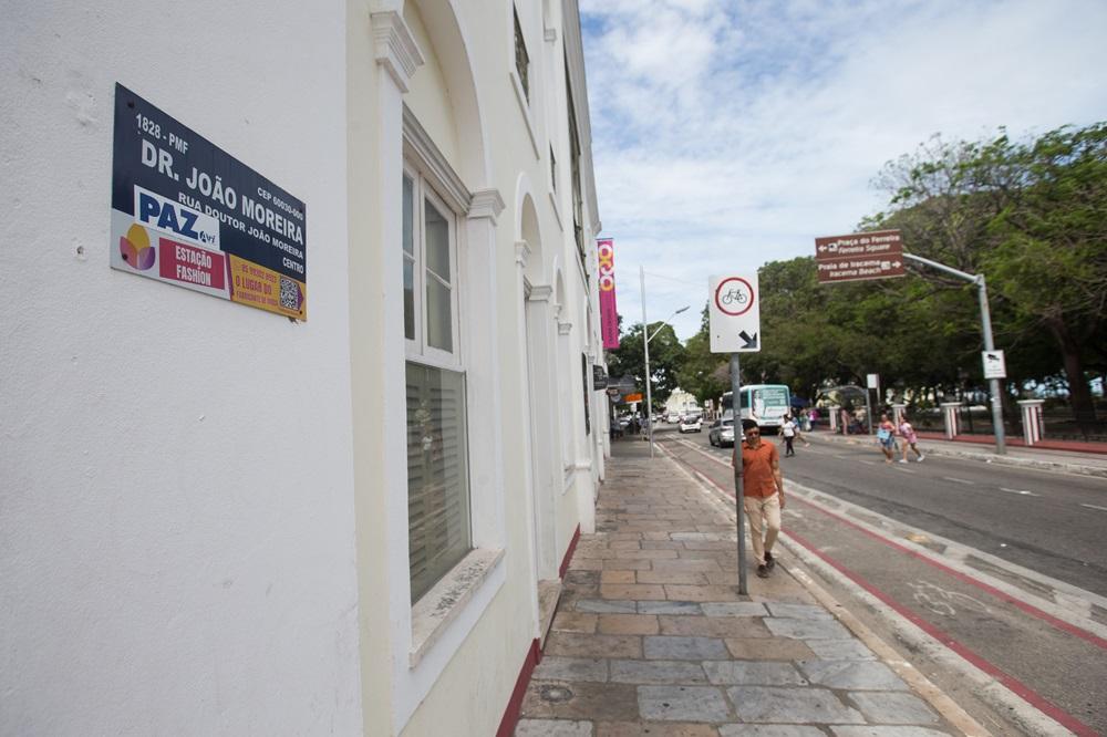 Na imagem, uma fotografia em perspectiva de uma calçada na Rua Doutor João Moreira, no centro de Fortaleza. No primeiro plano, à esquerda, destaca-se uma placa de identificação de logradouro afixada em uma parede branca de um edifício histórico. A placa contém o nome 