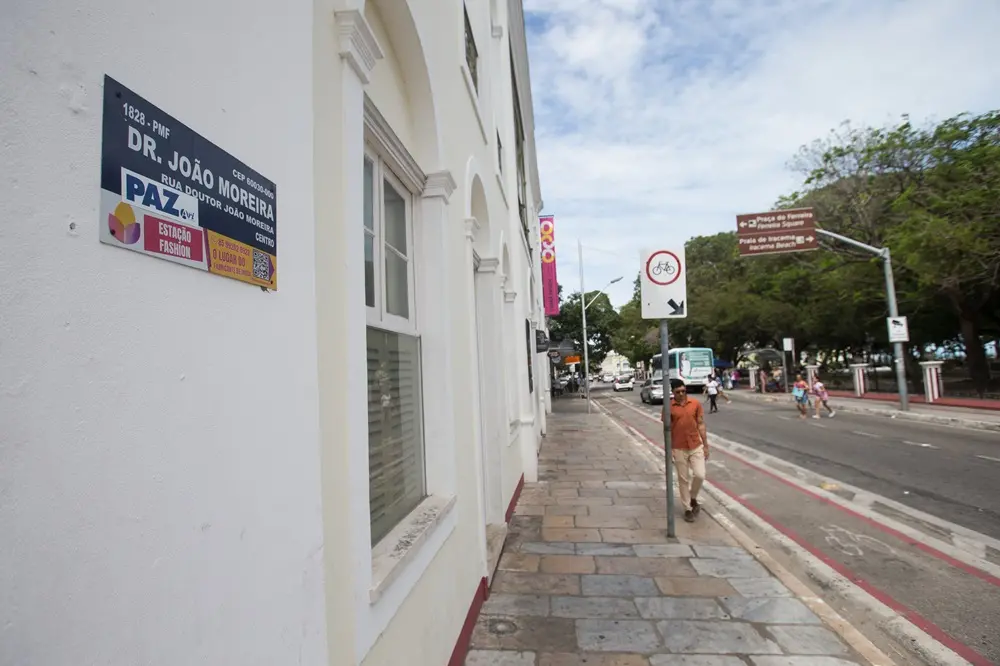 Na imagem, uma fotografia em perspectiva de uma calçada na Rua Doutor João Moreira, no centro de Fortaleza. No primeiro plano, à esquerda, destaca-se uma placa de identificação de logradouro afixada em uma parede branca de um edifício histórico. A placa contém o nome 