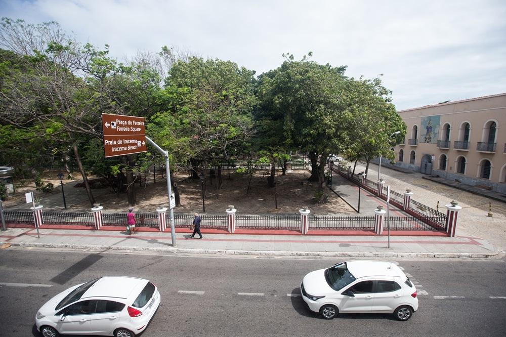 Na imagem, captura uma vista aérea de uma rua urbana em Fortaleza, Brasil. No centro da composição, há uma praça cercada por uma grade de metal preta com colunas brancas e detalhes em vermelho. A praça é densamente arborizada, com árvores de copas verdes e frondosas que projetam sombras sobre o solo de terra batida. À esquerda, uma placa de sinalização turística marrom indica as direções para a Praça do Ferreira e a Praia de Iracema. No primeiro plano, dois carros brancos circulam por uma avenida asfaltada com sinalização horizontal. Ao fundo, à direita, destaca-se um edifício histórico de dois andares em tom bege, com janelas em arco e um painel de azulejos na fachada. O céu está claro com poucas nuvens, sugerindo um dia ensolarado.