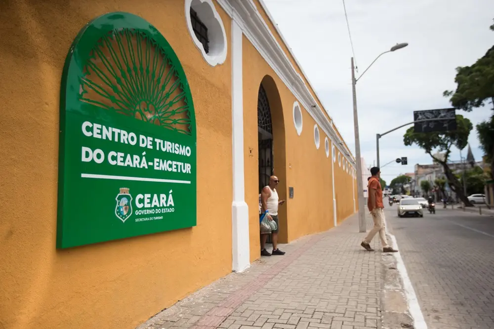 Na imagem, fotografia em plano médio da fachada lateral do Centro de Turismo do Ceará (Emcetur), em Fortaleza. O prédio histórico possui paredes pintadas de ocre amarelado com detalhes em branco. Em destaque, à esquerda, há uma grande placa verde com o logotipo do Governo do Estado do Ceará. Na calçada de pedras portuguesas, um homem de camiseta branca observa o movimento, enquanto outro homem, vestindo camisa laranja, caminha em direção à rua. Ao fundo, vê-se o fluxo de carros e a arborização da Rua João Moreira sob um céu claro.