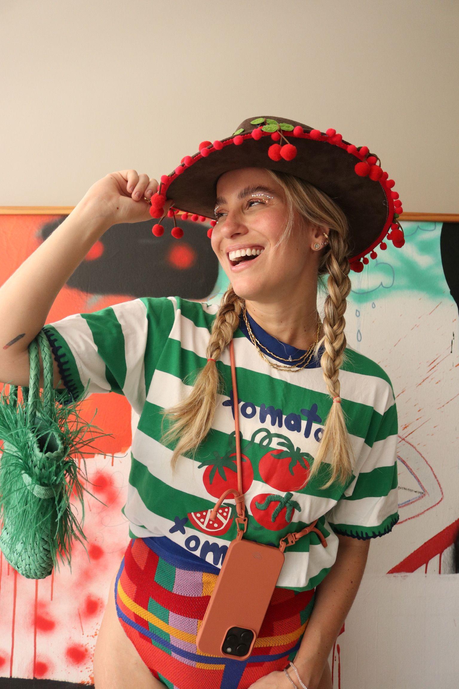 Jovem mulher sorrindo com tranças, usando um chapéu com pompons vermelhos e uma camiseta listrada com estampa de tomate, segurando uma bolsa verde texturizada.