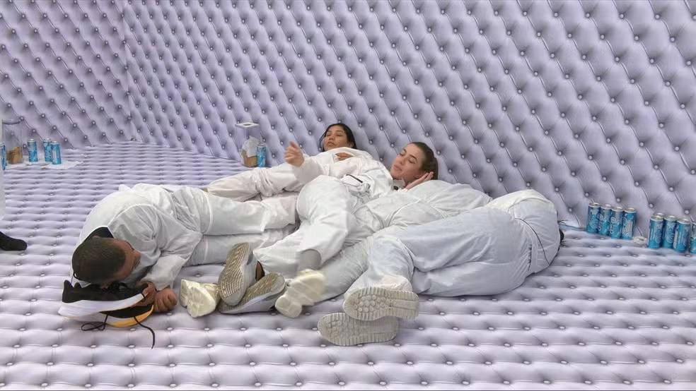 Participantes do reality show Big Brother Brasil estão deitados e amontoados no chão do Quarto Branco, todos vestindo macacões brancos e cercados por paredes. O ambiente monocromático é quebrado apenas pelas latas de bebida azuis espalhadas ao redor e pelos tênis dos integrantes em destaque no primeiro plano.