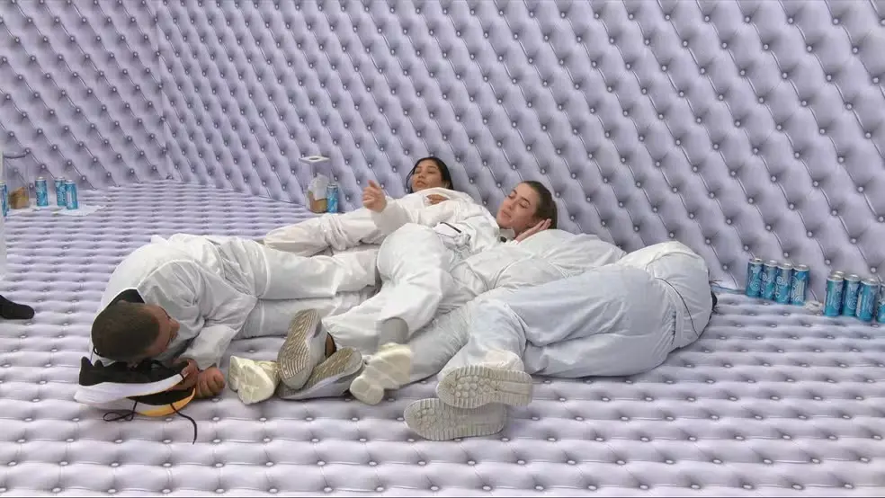 Participantes do reality show Big Brother Brasil estão deitados e amontoados no chão do Quarto Branco, todos vestindo macacões brancos e cercados por paredes. O ambiente monocromático é quebrado apenas pelas latas de bebida azuis espalhadas ao redor e pelos tênis dos integrantes em destaque no primeiro plano.
