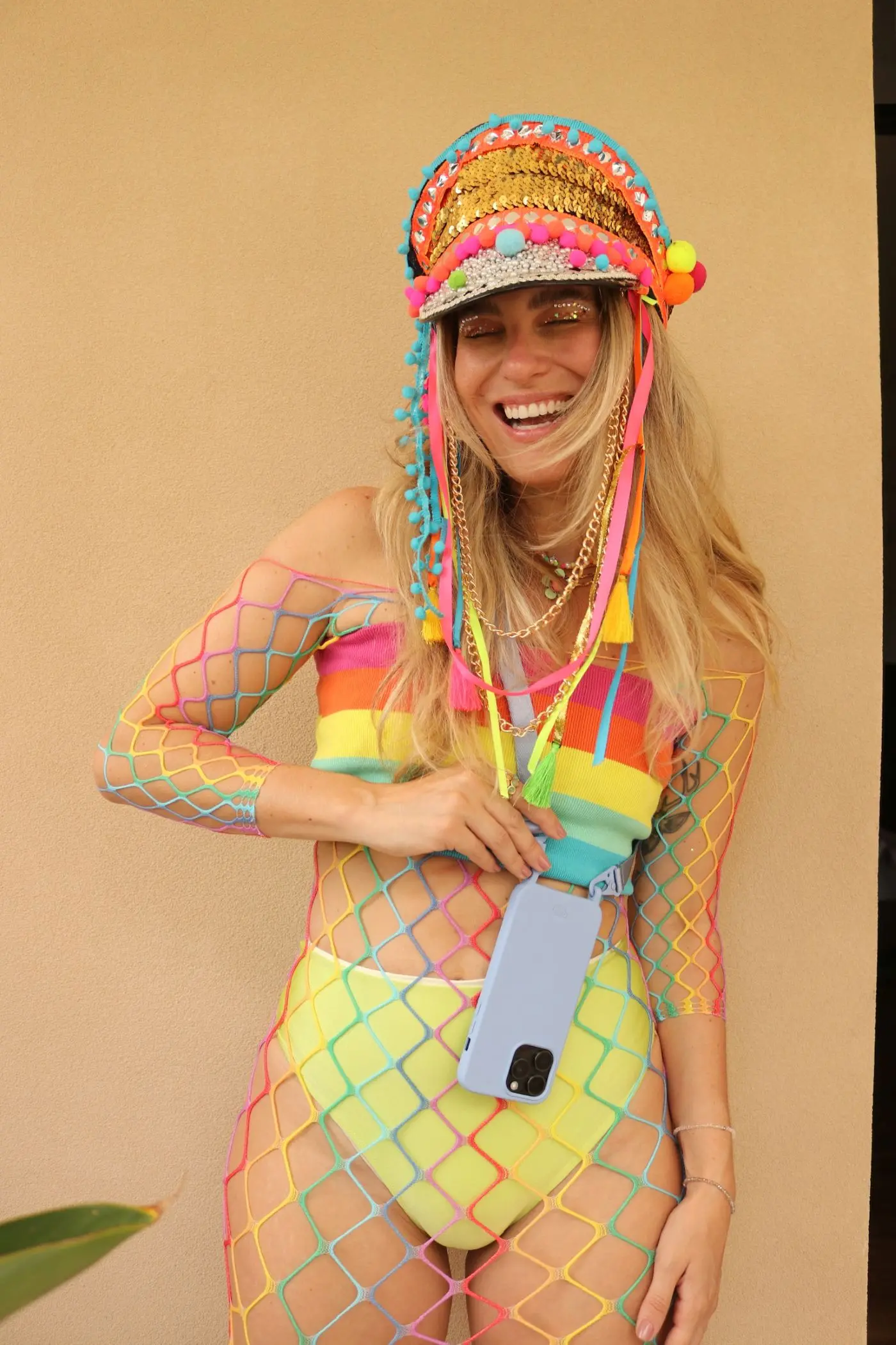 Mulher loira sorridente vestindo um chapéu de crochê colorido, um body de rede vibrante e um biquíni amarelo neon, segurando um smartphone.