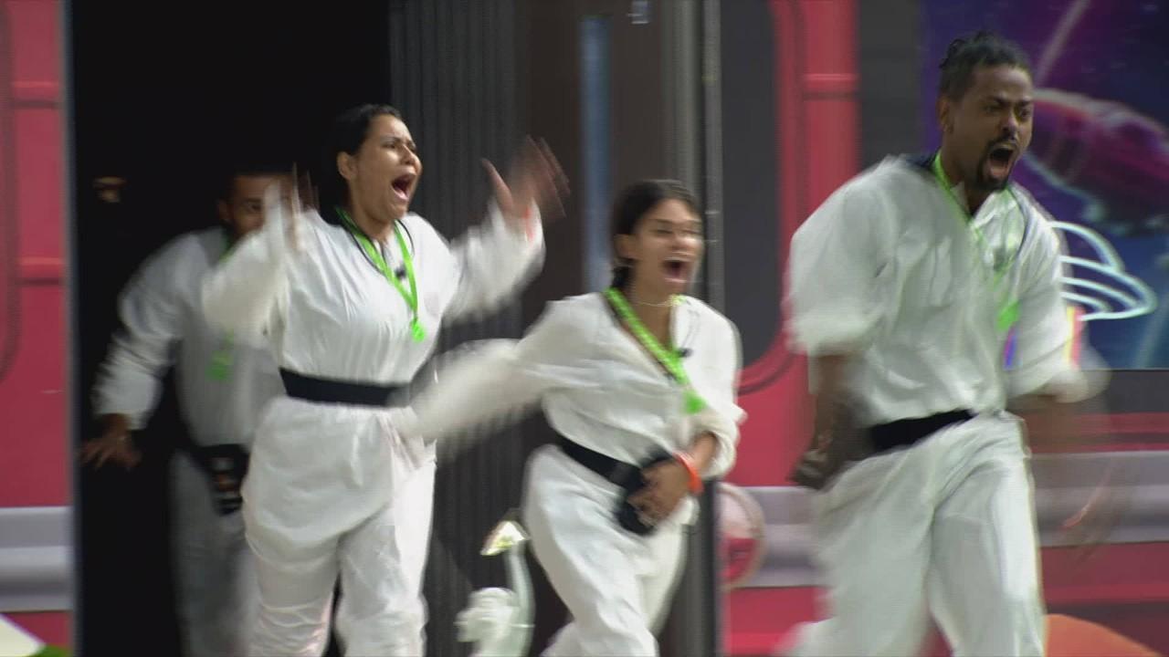 Participantes do BBB 26 correm e gritam entusiasmados ao entrar na casa, vestindo macacões brancos com faixas pretas e cordões verdes. A cena captura um momento de intensa euforia e movimento, com os integrantes saindo do quarto branco para o cenário da casa do reality.
